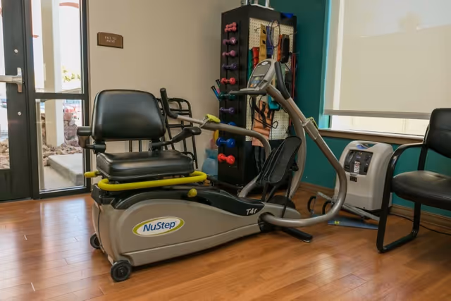 A small exercise room with a NuStep T4r recumbent cross trainer, a rack of colorful dumbbells and resistance bands, a black chair, and a window with a white roller shade partially down.