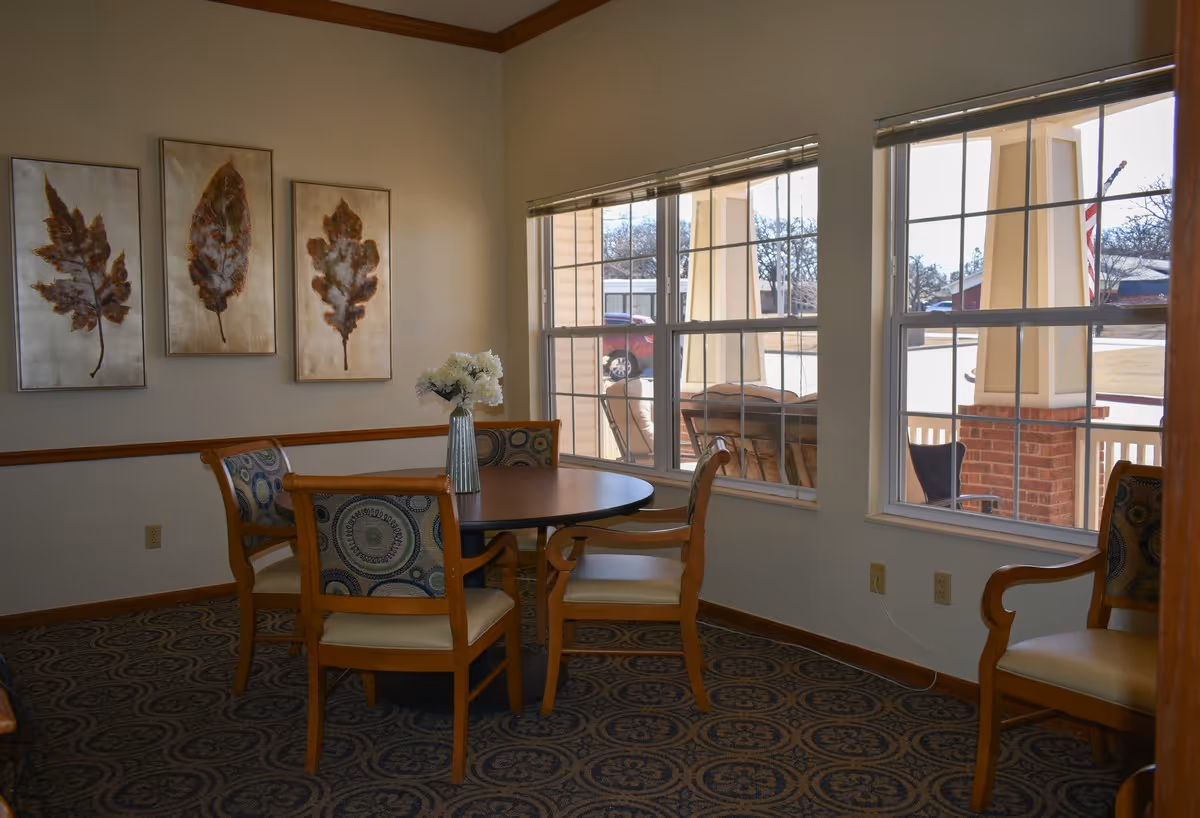 A small dining area with a round table, four chairs, a vase of flowers, leaf artwork on the wall, and large windows looking out to a porch.