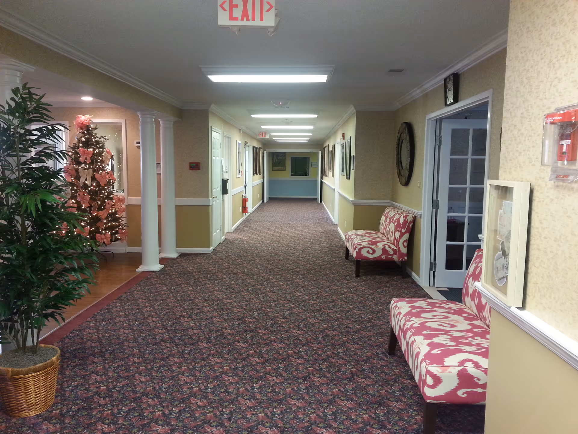 A long, carpeted hallway in a senior living facility with floral-patterned benches along the right wall and a decorated Christmas tree with pink bows on the left. The hallway is well-lit with ceiling lights and has several framed pictures on the walls. There is an exit sign hanging from the ceiling at the far end of the hallway.