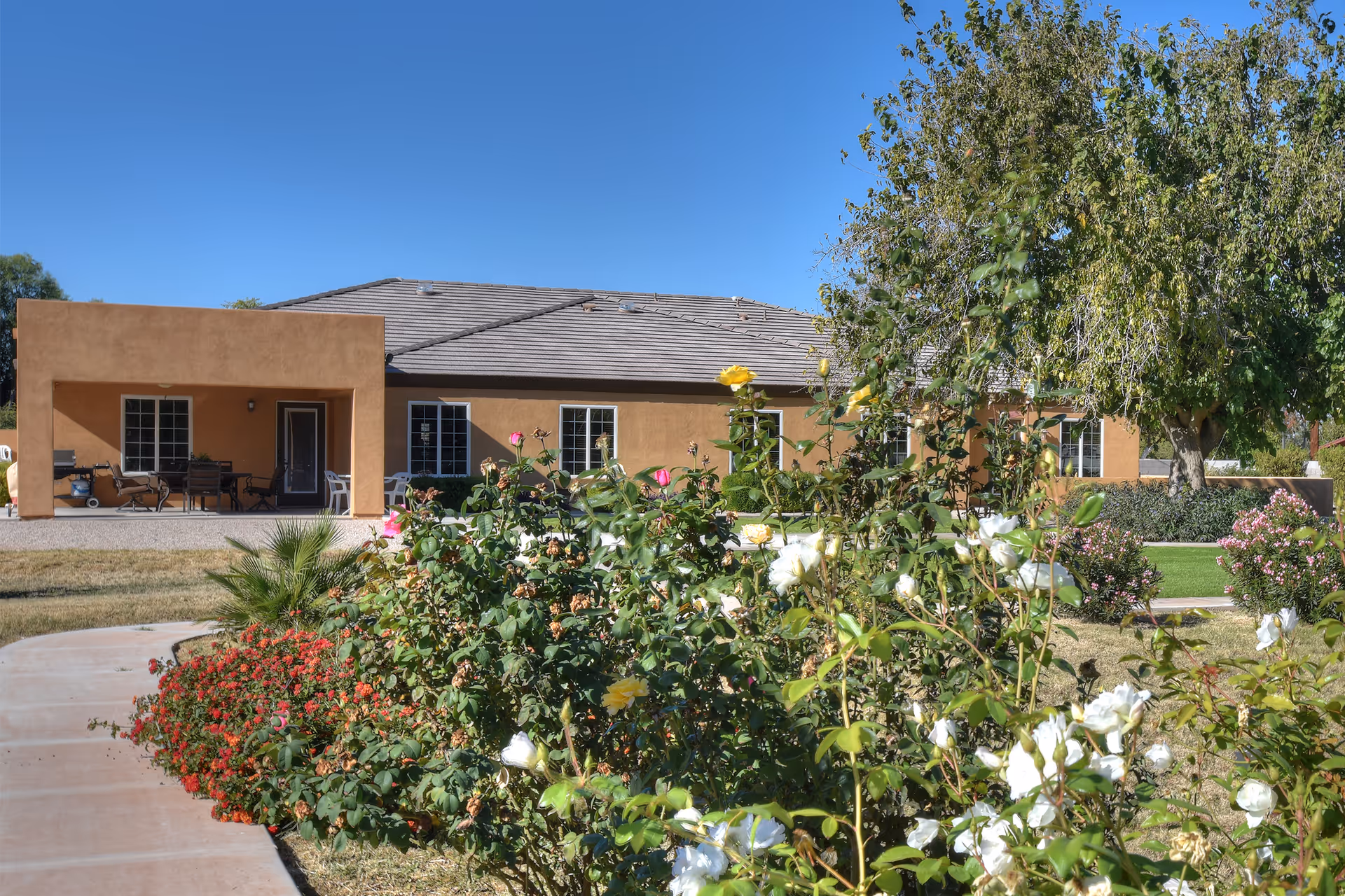A single-story building with a tan exterior and a gray tiled roof, surrounded by a garden with various flowering plants and bushes. A paved walkway curves through the garden leading to the building's entrance. The sky is clear and blue.