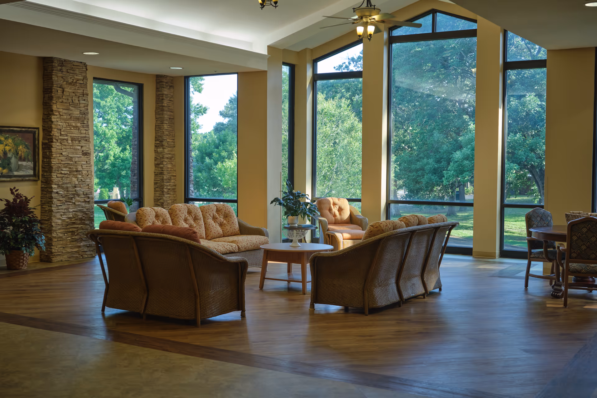 A spacious living room area with large floor-to-ceiling windows showing a green outdoor view. The room features wicker furniture with cushioned seats arranged around a small round wooden table. There are stone accent walls, a ceiling fan with lights, and a wooden floor.