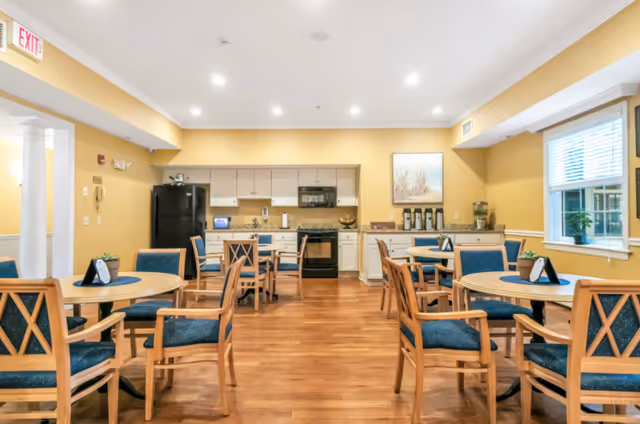 A bright and clean dining area in a senior living facility with round wooden tables and blue cushioned chairs. The room has yellow walls, wood flooring, and a kitchen area in the background with white cabinets, a black refrigerator, stove, and microwave. There are small plants and table decorations on the tables, and a window with blinds on the right side.