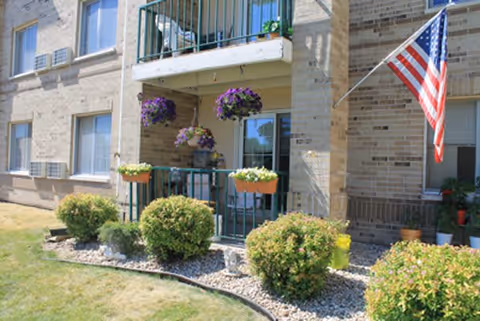 Ground-floor porch and balcony on a brick building with hanging flower baskets, potted plants, an American flag, and landscaped shrubs and rock beds.
