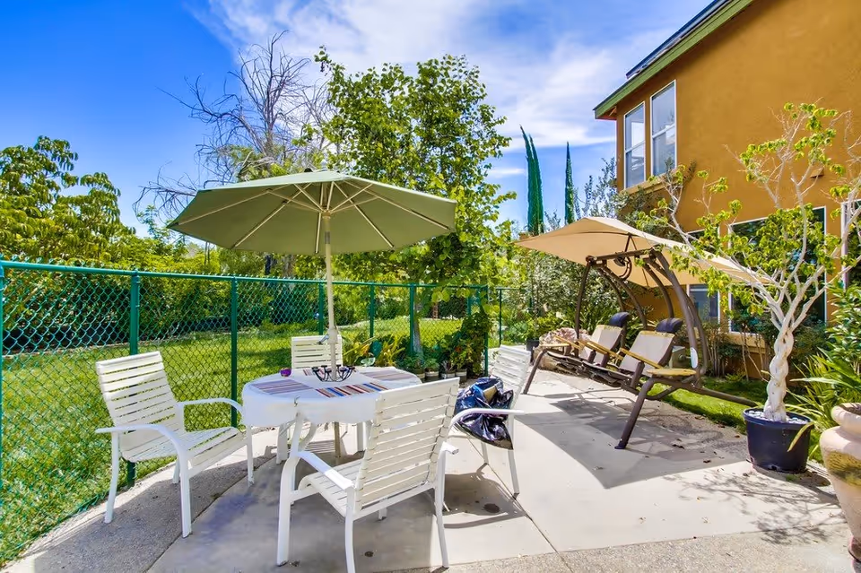 Outdoor patio area with a round table covered by a green umbrella surrounded by four white chairs. To the right, there is a beige cushioned swing with a canopy. The patio is adjacent to a yellow building and enclosed by a green chain-link fence with trees and greenery in the background under a blue sky.