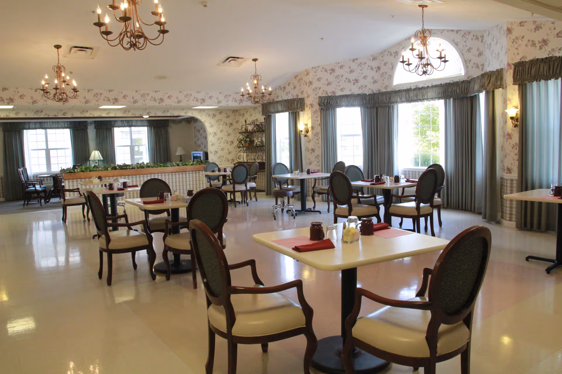 A bright dining room with several tables and chairs arranged neatly. The room features floral wallpaper, large windows with curtains, and chandeliers hanging from the ceiling. Tables are set with red napkins, salt and pepper shakers, and small pitchers.