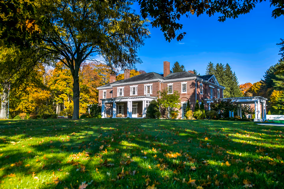 Large two-story brick mansion front with a wide lawn and trees showing autumn foliage under a clear blue sky.