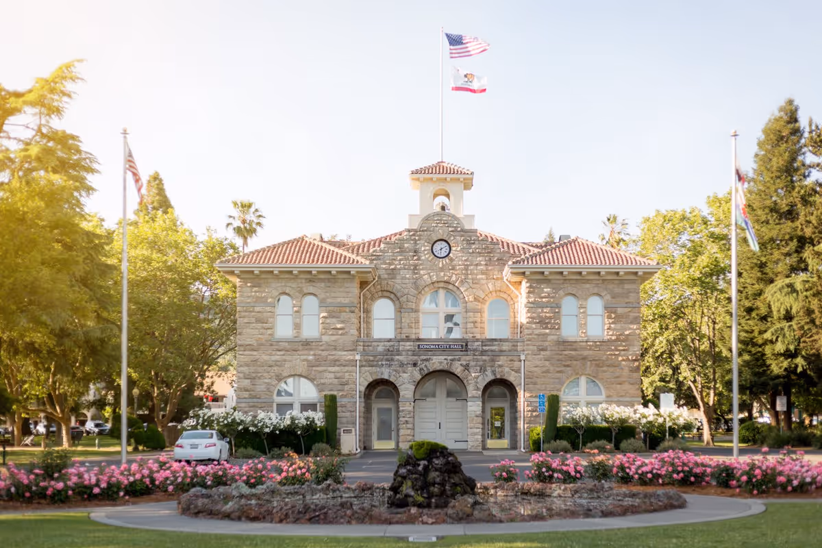 Stone building with a red tile roof and a clock above the main entrance, identified as Sonoma City Hall, surrounded by trees, flower beds with pink flowers, and three flagpoles with flags flying.