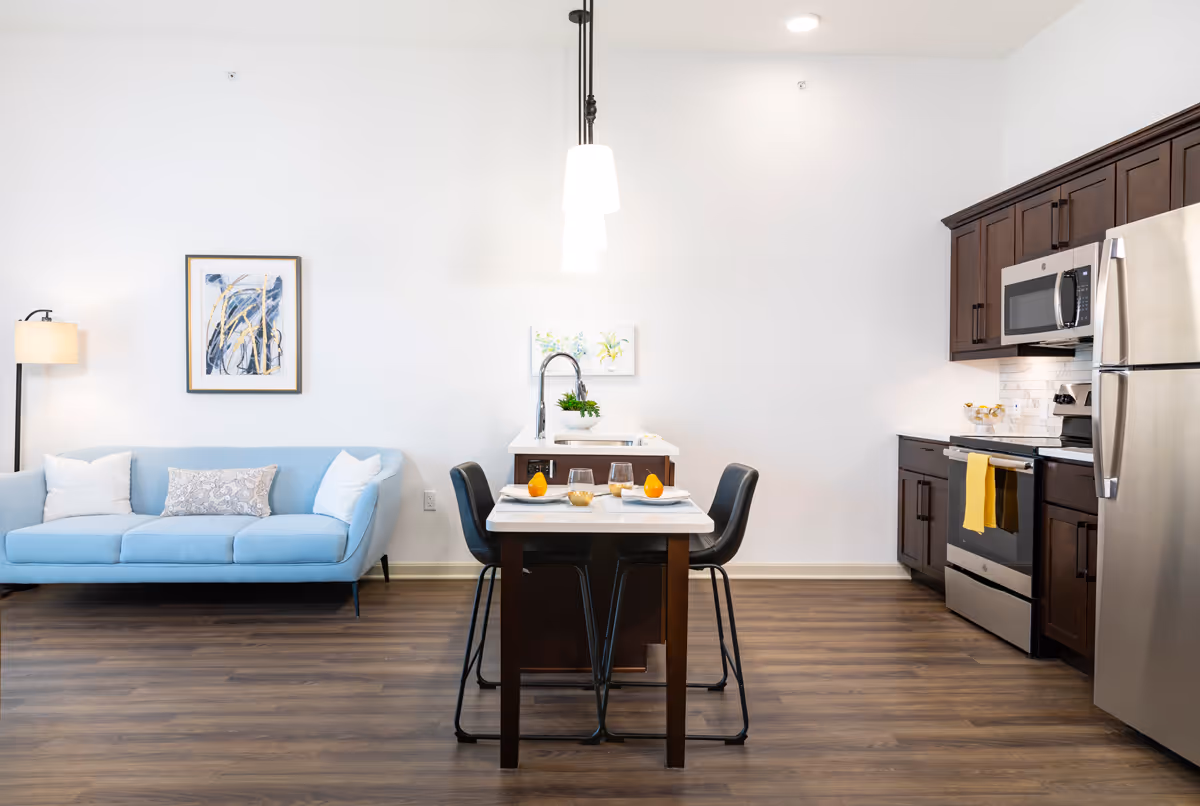 A modern open-concept living and kitchen area featuring a light blue sofa with three pillows on the left, a small dining table with four black chairs in the center, and a kitchen with dark wood cabinets, stainless steel appliances including a refrigerator, microwave, and stove on the right. The room has wooden flooring, white walls, two hanging pendant lights above the dining table, and framed artwork on the walls.