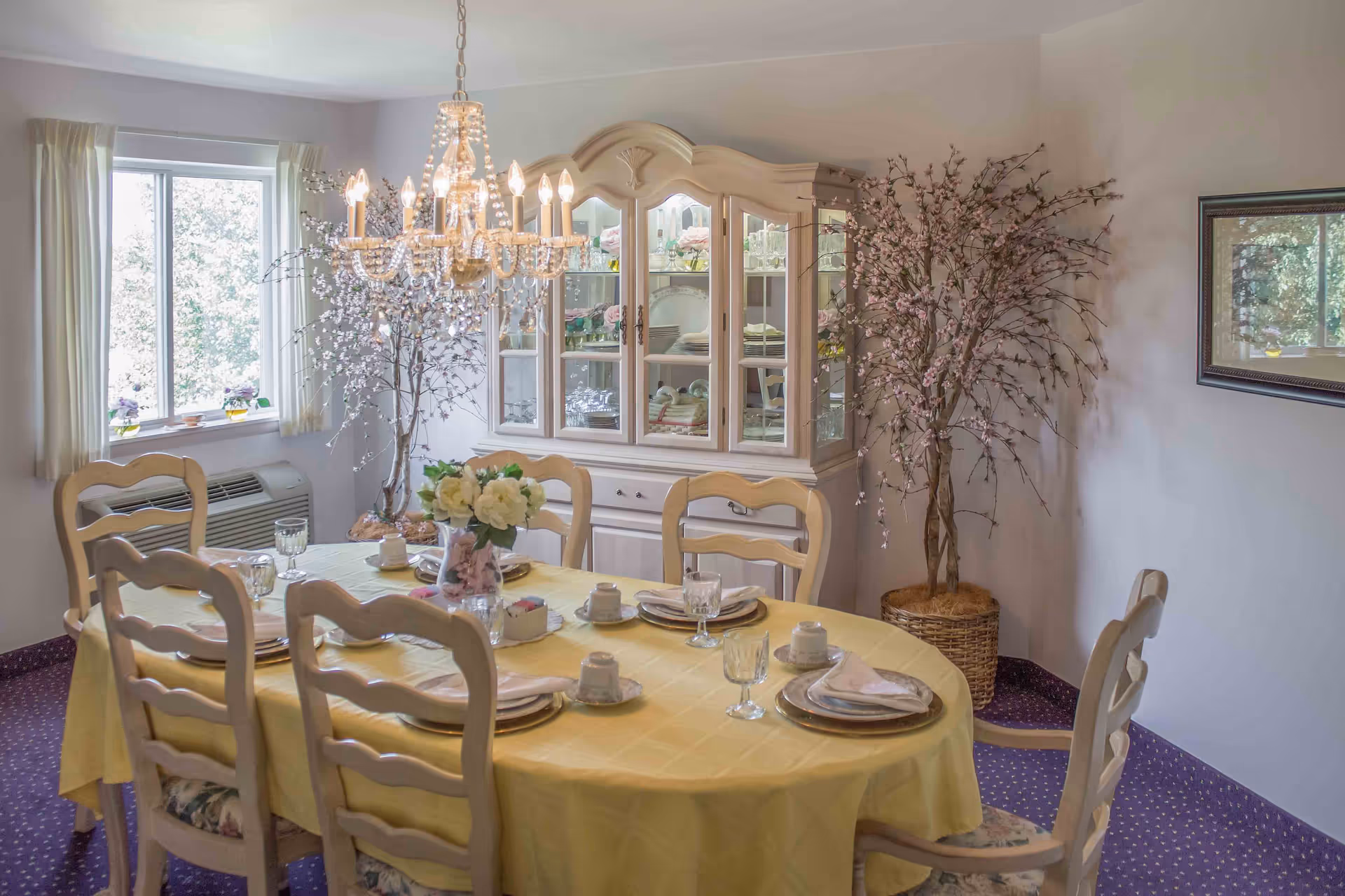 Dining room with a chandelier and a table set for six covered by a yellow tablecloth, a glass-front china cabinet, and potted decorative branches.