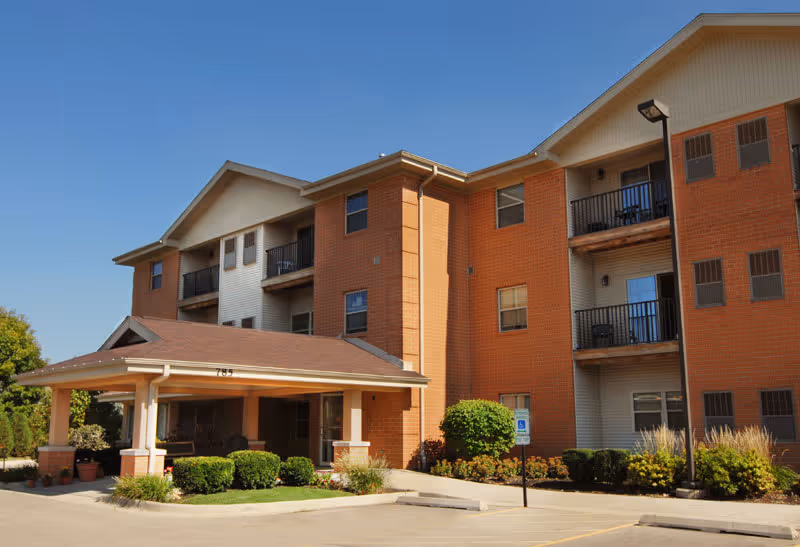 Exterior view of a three-story senior living facility building with a covered entrance, brick and siding facade, balconies, and landscaped bushes under a clear blue sky.