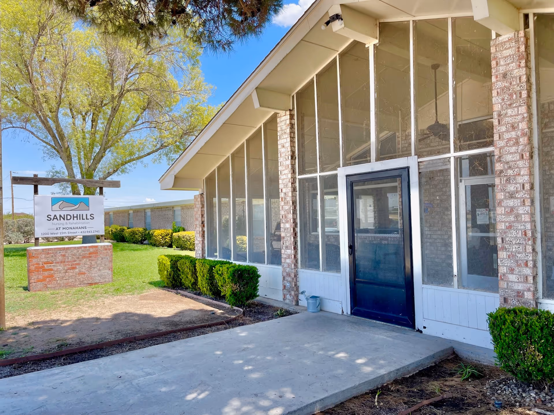 Front entrance of the Sandhills Nursing and Rehabilitation building with large screened windows, a glass door, brick facade, and a lawn sign.