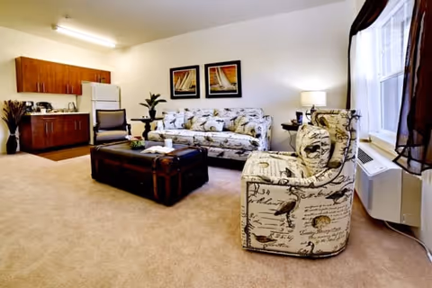 A cozy living room area with a patterned sofa and matching armchair, a dark wooden coffee table, a single chair, and a small kitchenette with wooden cabinets, a refrigerator, and a microwave. The room has beige carpet, white walls, two framed pictures above the sofa, a window with dark curtains, and a lamp on a side table.