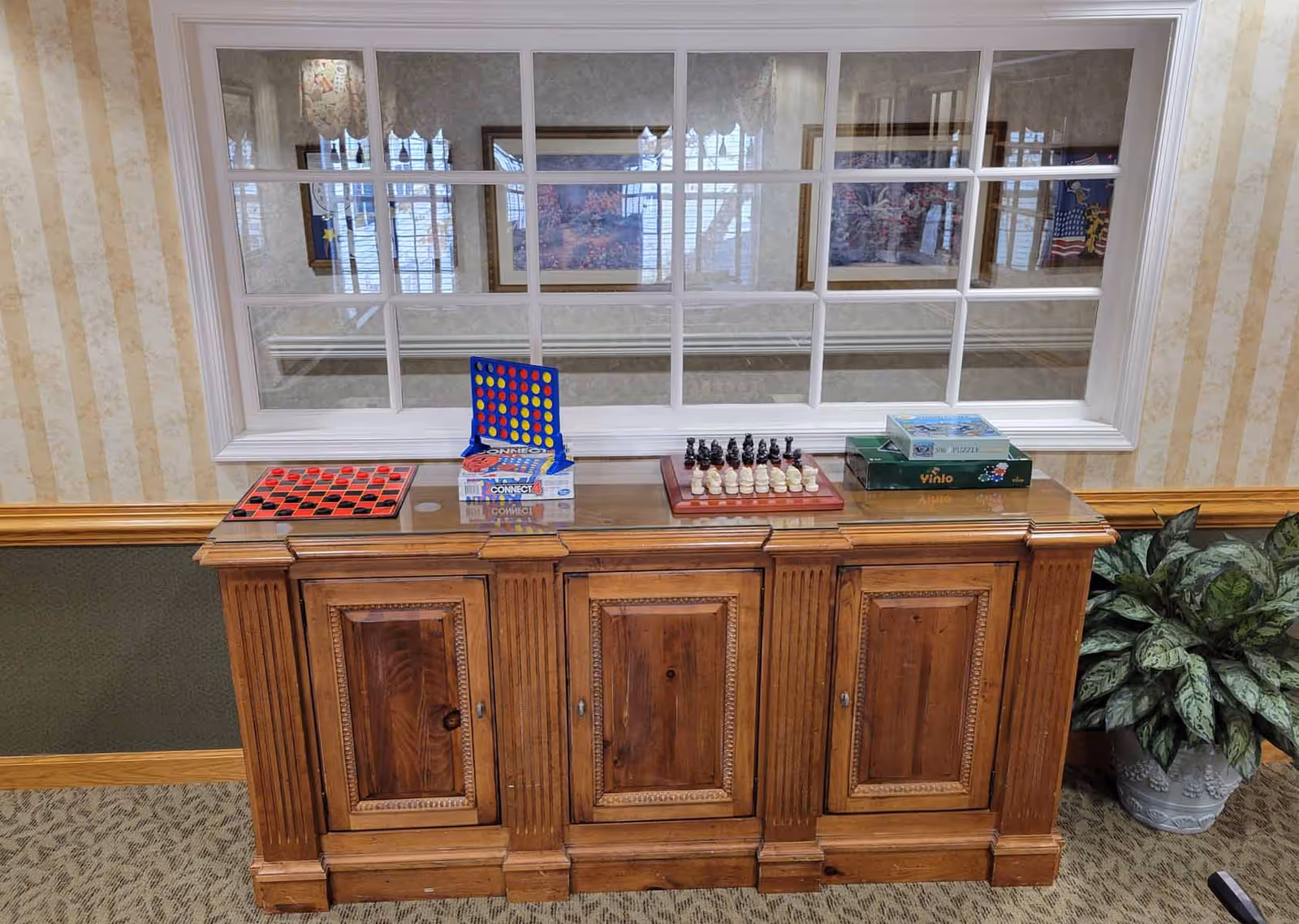 A wooden cabinet with a glass top holding various board games including checkers, Connect 4, chess, and puzzles. Behind the cabinet is a large window with multiple panes, reflecting framed artwork and windows from the opposite side. To the right of the cabinet is a potted plant with green and white leaves. The room has patterned wallpaper and carpeted flooring.