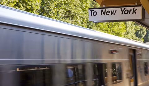 A blurred subway train passing a platform sign that reads "To New York" with trees in the background.