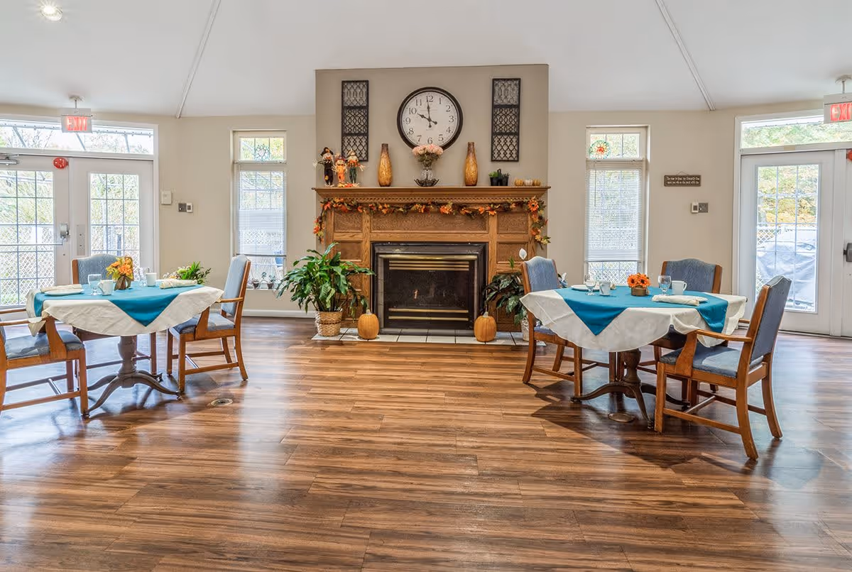A cozy dining area with two round tables covered with white and teal tablecloths, each surrounded by four wooden chairs with blue cushions. The room features a wooden floor, a central fireplace decorated with autumn-themed garlands, pumpkins, and plants. Above the fireplace is a large clock and decorative wall hangings. Large windows and glass doors allow natural light to fill the space.