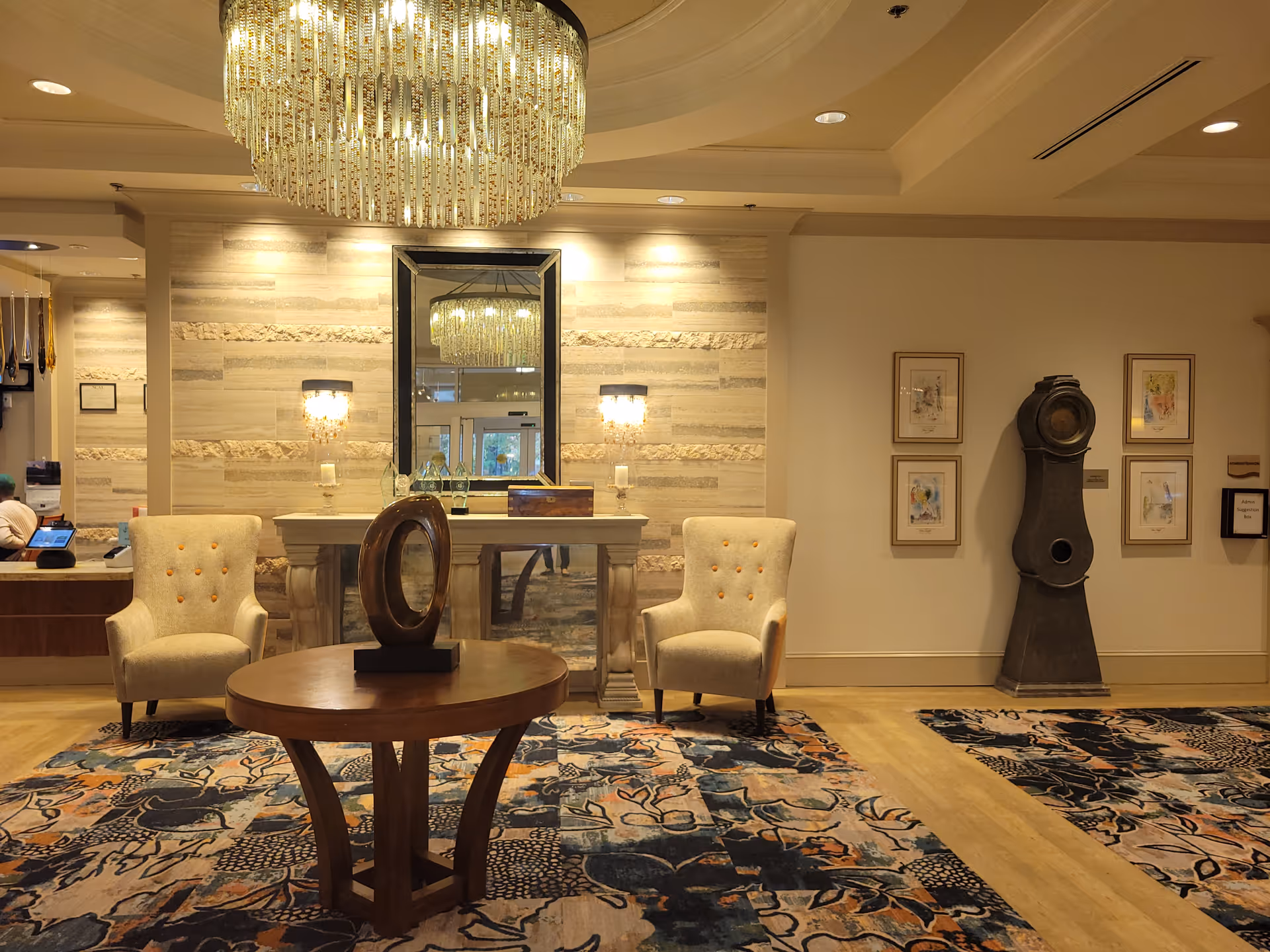 A well-lit interior lobby area with a large chandelier hanging from the ceiling. There is a round wooden table with a decorative sculpture in the center, flanked by two beige armchairs with orange buttons. Behind the table is a console table with a large mirror and two wall sconces on either side. The floor is covered with a patterned carpet, and there is a tall, dark grandfather clock against the wall with framed artwork surrounding it.