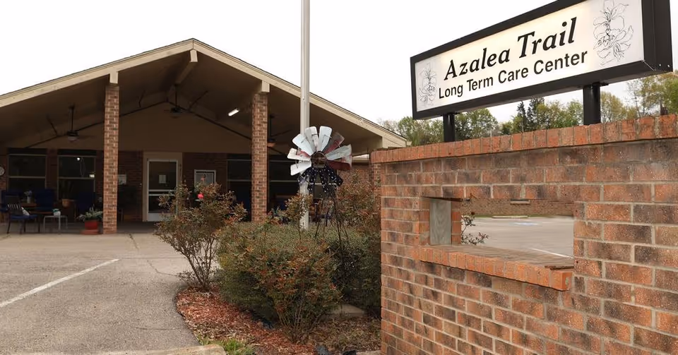 Exterior view of Azalea Trail Long Term Care Center showing the entrance with a covered porch supported by brick pillars, some outdoor seating, a small garden with bushes and a decorative windmill, and a brick sign with the facility name.