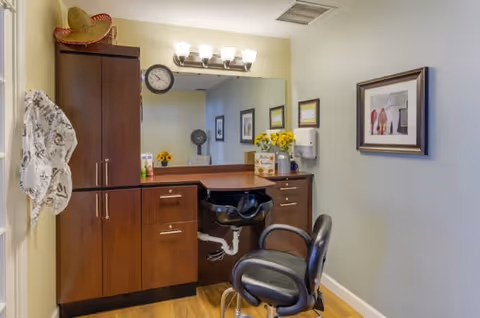 Interior view of a small salon or grooming area with a black salon chair in front of a wooden vanity with a mirror and overhead lights. The vanity has cabinets and drawers, with a clock on the wall reflected in the mirror. There are decorative items including a vase with yellow flowers and framed pictures on the walls. A hat hangs on the left wall.