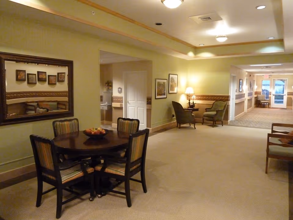 Interior view of a senior living facility hallway with a round wooden table and four striped chairs in the foreground. A large mirror hangs on the wall above the table. In the background, there are two armchairs with a small table and lamp between them, framed pictures on the walls, and a hallway leading to a door with windows.