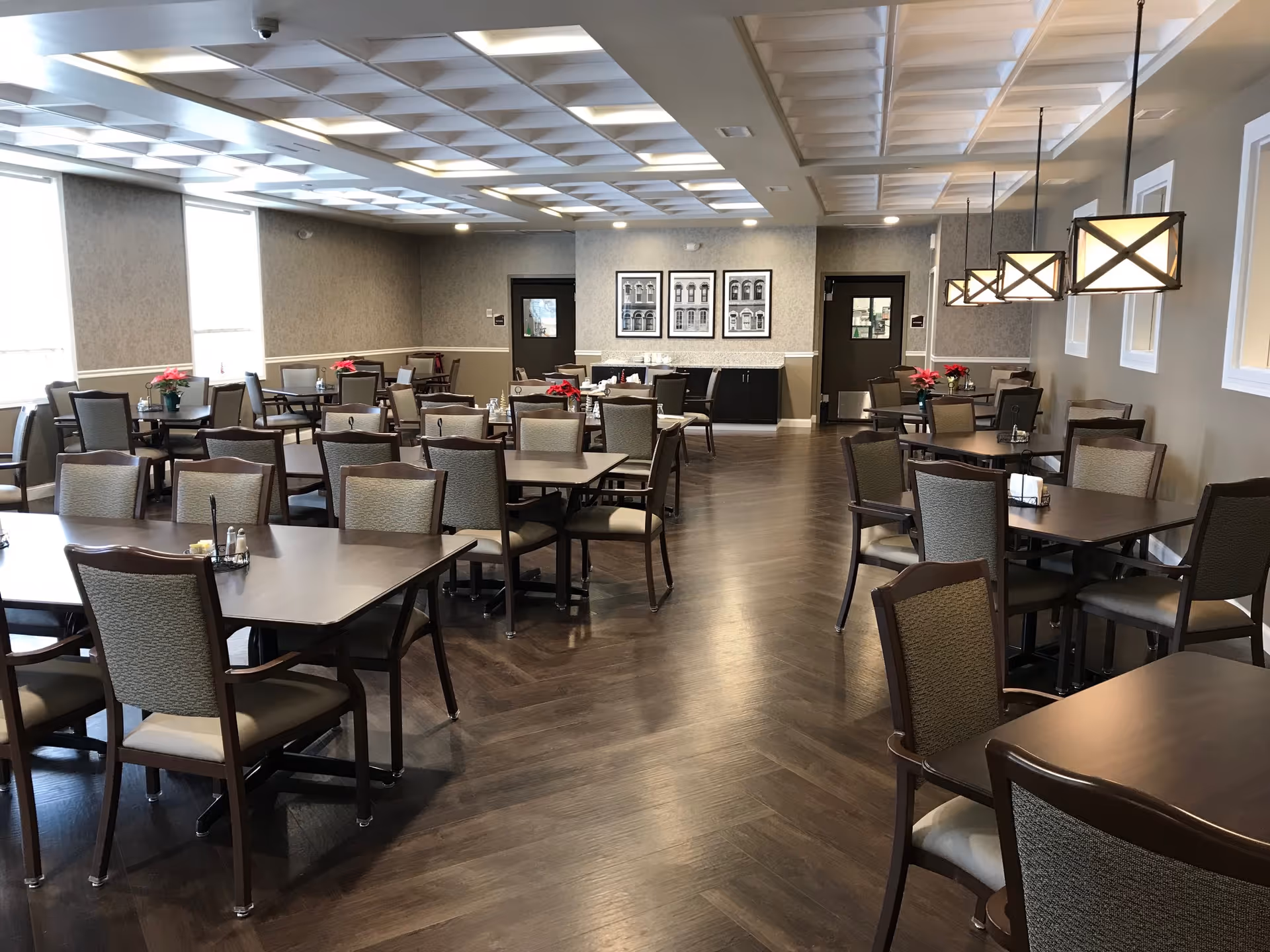 A spacious dining room with multiple dark wood tables and cushioned chairs arranged neatly. The room has large windows on the left side allowing natural light, modern pendant lights hanging from the ceiling on the right, and framed black and white artwork on the far wall. The floor is a dark wood pattern, and small flower arrangements are placed on some tables.