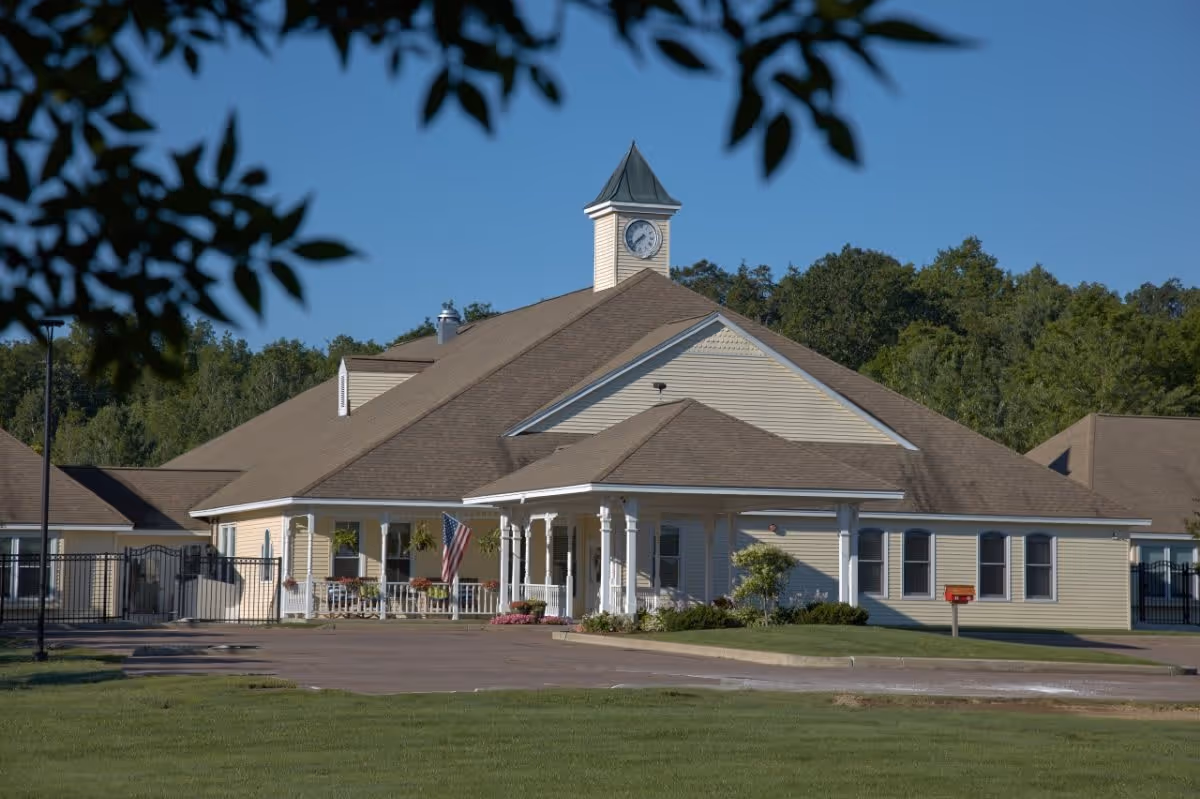 Exterior view of a single-story building with beige siding and a brown roof, featuring a clock tower on top. The building has a covered porch with white columns, an American flag, and some plants. The surrounding area includes a paved driveway, green lawn, and trees in the background under a clear blue sky.