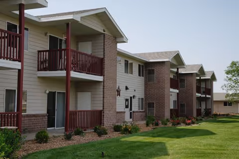 Exterior view of a two-story residential building with balconies and a well-maintained lawn and garden beds in front. The building features beige siding with brick accents and red railings on the balconies.