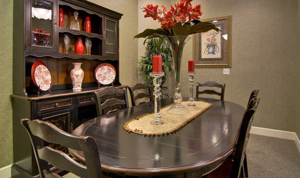 A dining room with a dark wooden oval table surrounded by six matching chairs. On the table is a beige runner with three glass candle holders holding red candles and a tall glass vase with red flowers. Behind the table is a dark wooden hutch displaying decorative plates, vases, and glassware. The walls are light green with a framed floral painting and a potted plant in the corner.