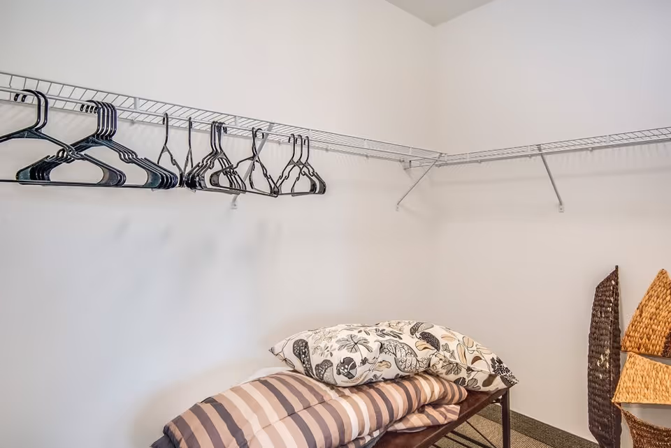 Small walk-in closet with wire shelving and empty hangers, a bench holding folded bedding and pillows, and wicker baskets in the corner.