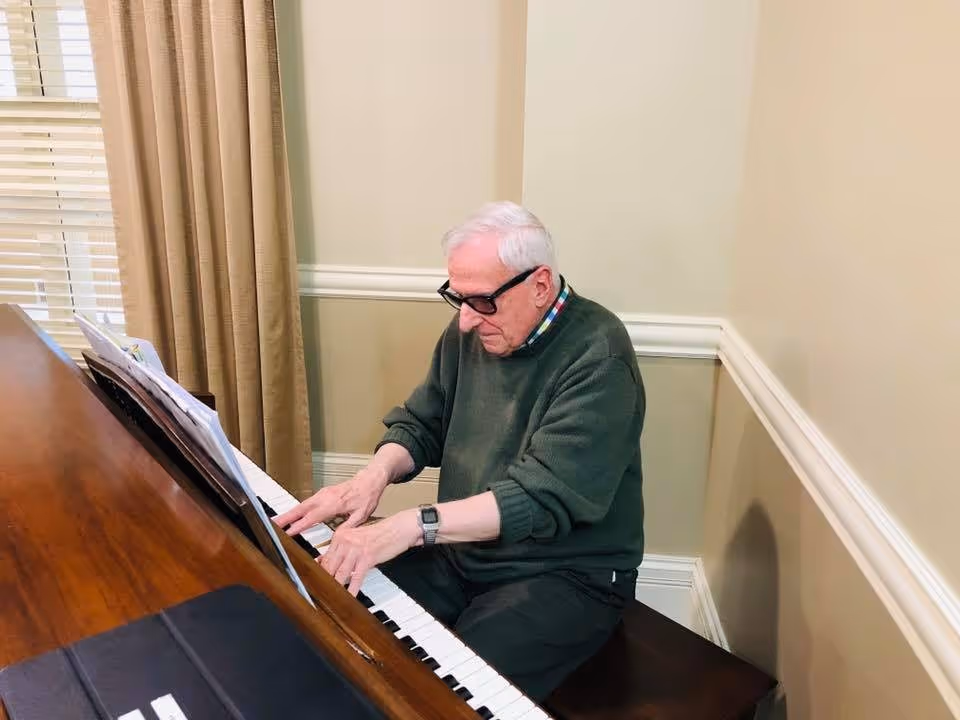 An elderly man wearing glasses and a green sweater is playing a wooden piano in a room with beige walls and a window with beige curtains.