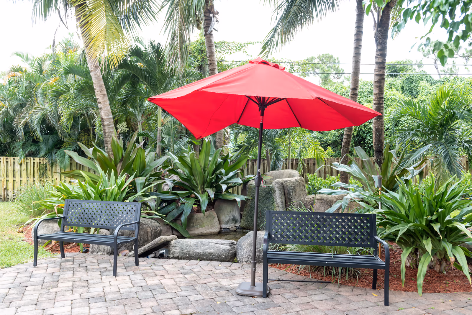 Outdoor patio area with two black metal benches and a large red umbrella in the center. The area is paved with stone tiles and surrounded by lush green plants, palm trees, and a wooden fence in the background.