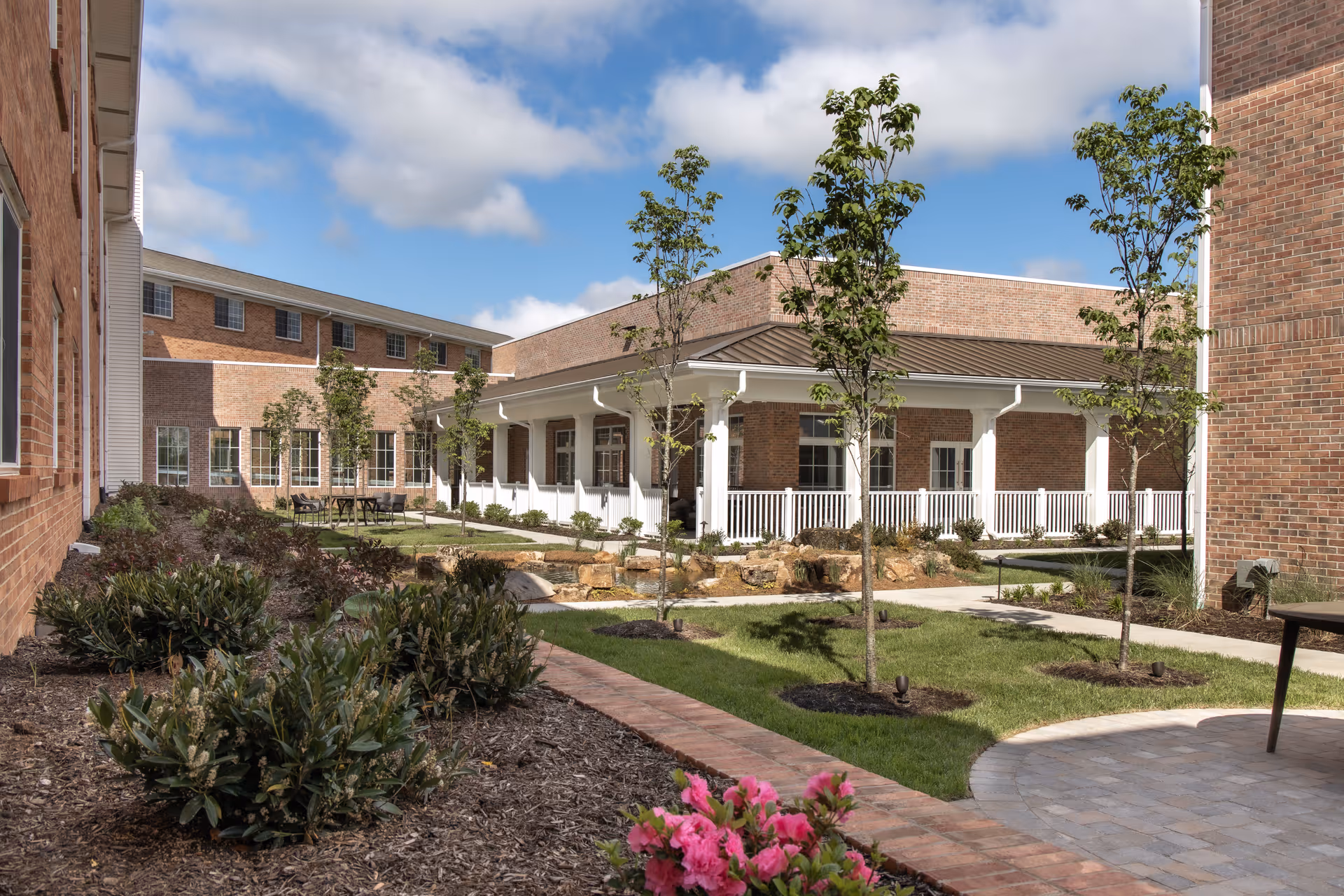 Outdoor courtyard area of a senior living facility with brick buildings surrounding a landscaped garden. The garden features small trees, shrubs, a paved walkway, and a covered patio with white railings. The sky is partly cloudy with blue patches visible.