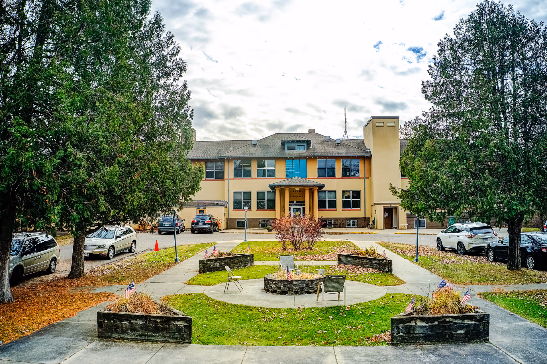 Front exterior view of a two-story building with a beige and brown facade, surrounded by large trees and a parking area with several cars. In the foreground, there is a circular paved area with chairs around a fire pit, small garden beds with American flags, and walkways leading to the building entrance.