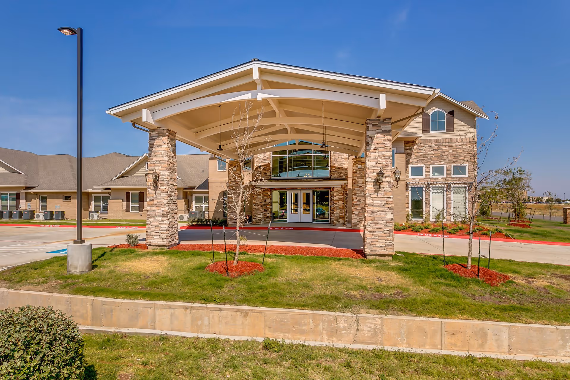 Front exterior view of Bristol Park at Amarillo Assisted & Living Memory Care facility showing a covered entrance supported by stone pillars, landscaped grass and small trees, and a clear blue sky.