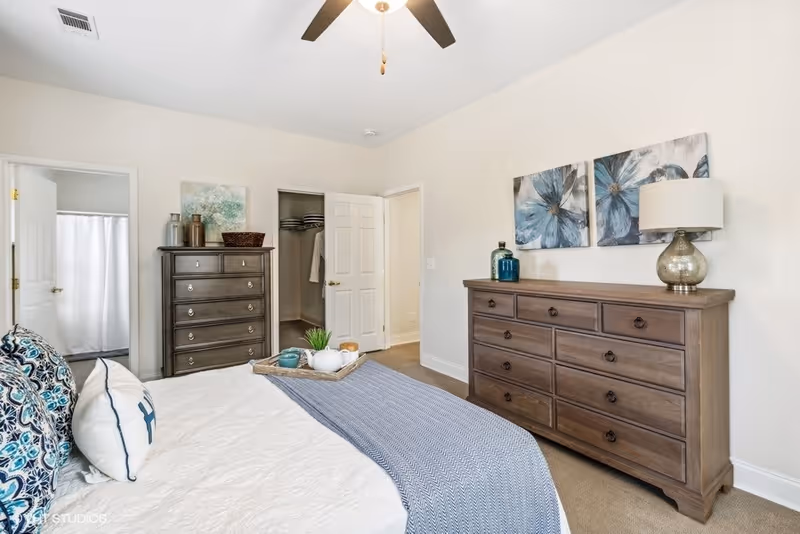 A bright and neatly arranged bedroom featuring a bed with patterned pillows and a blue throw blanket. There are two wooden dressers, one tall and one wide, with decorative items including a lamp and artwork on the walls. The room has a ceiling fan and an open door leading to a walk-in closet and a bathroom with a shower curtain.