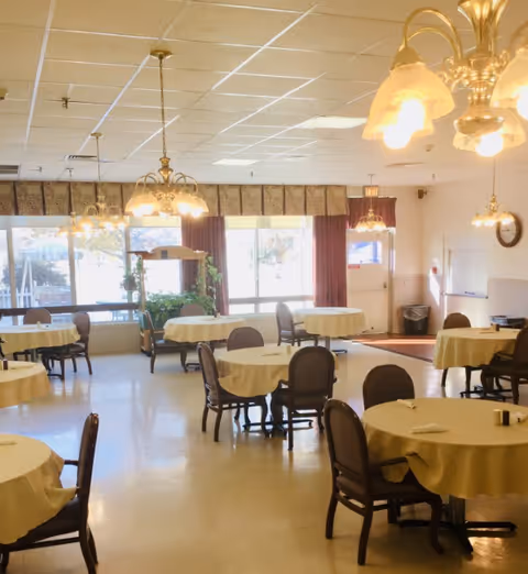 A bright dining room with multiple round tables covered with yellow tablecloths and surrounded by chairs. The room has large windows with curtains allowing natural light to enter, and several hanging light fixtures with warm lighting. There is a clock on the wall and a trash bin near the door.