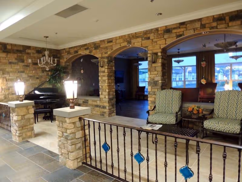 Indoor common lounge with stone archways, a grand piano, upholstered chairs, decorative iron railing and lamp posts.