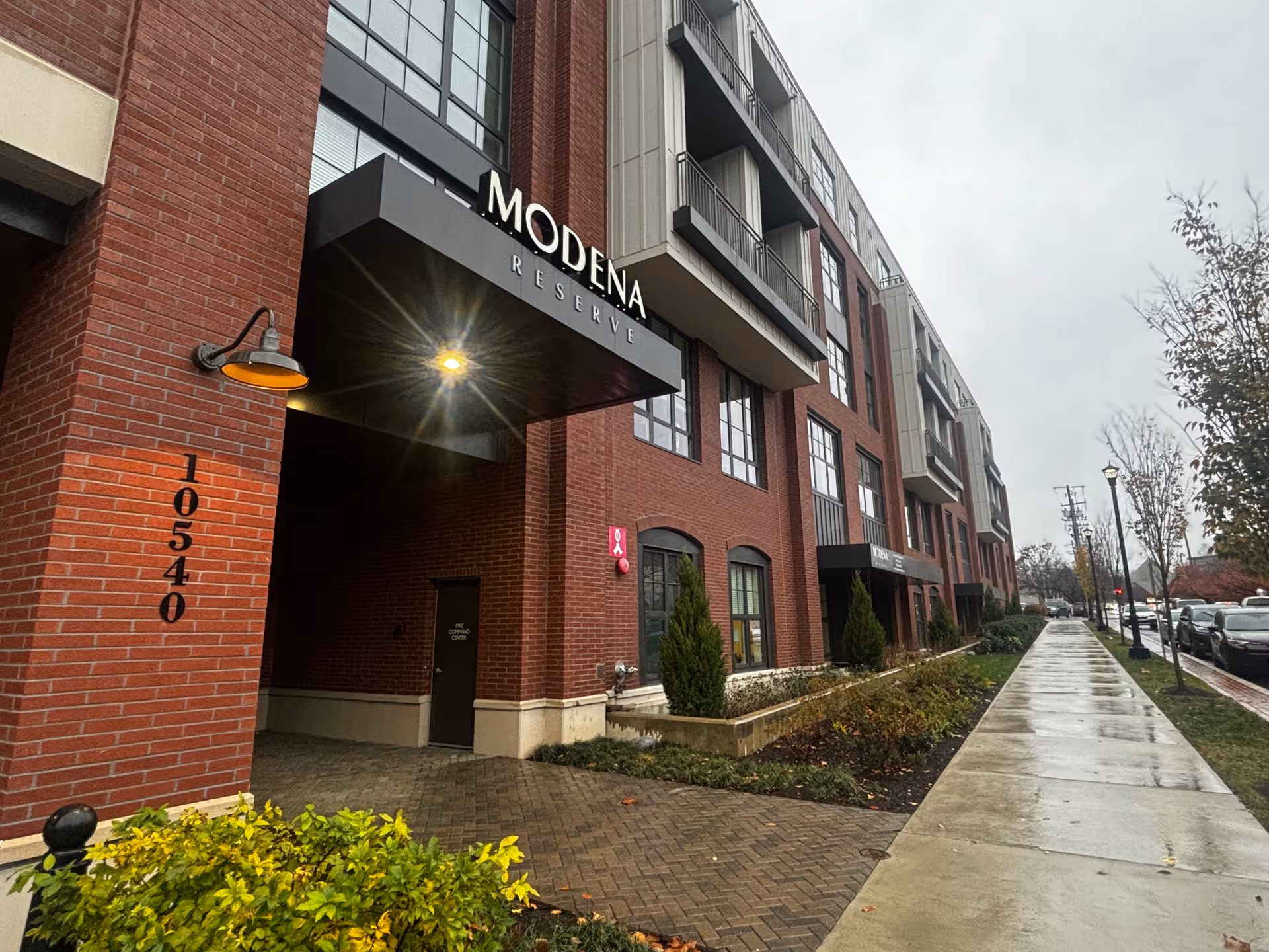 Exterior view of the Modena Reserve at Kensington building on a cloudy day, showing a brick facade with large windows, a covered entrance with the building name, and a wet sidewalk lined with small trees and parked cars.
