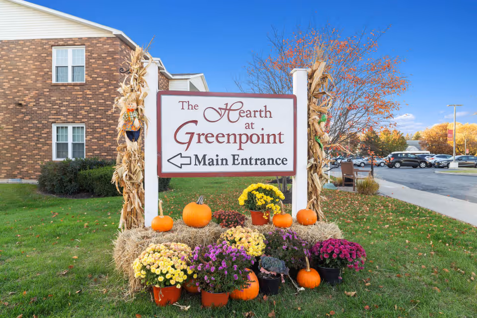 Outdoor sign for The Hearth at Greenpoint indicating the main entrance, surrounded by autumn decorations including pumpkins, hay bales, colorful potted flowers, and corn stalks tied to the signposts. A brick building and parked cars are visible in the background under a clear blue sky.