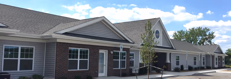 Exterior view of a single-story senior living facility building with gray siding and brick accents, multiple windows, a main entrance with a small ramp, and a clear blue sky with some clouds in the background.