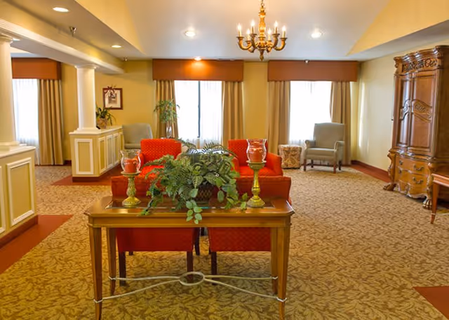 A cozy living room area with patterned carpet, two red armchairs, a wooden table with green plants and candle holders, two gray armchairs near windows with beige curtains, a wooden armoire, and a chandelier hanging from the ceiling.