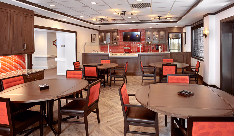 A dining room with several round wooden tables and chairs with red cushioned backs and seats. The room features a kitchen area in the background with dark wood cabinets, a red tiled backsplash, and various kitchen appliances. The floor has a light wood pattern, and the ceiling has recessed lighting and a decorative light fixture.