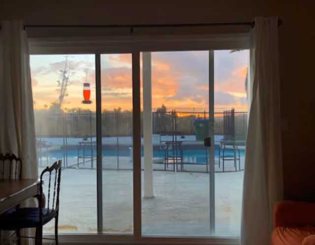 View through a sliding glass door showing an outdoor swimming pool area with a safety fence around it during sunset. Inside, part of a dining table with chairs is visible on the left and a portion of an orange sofa on the right.