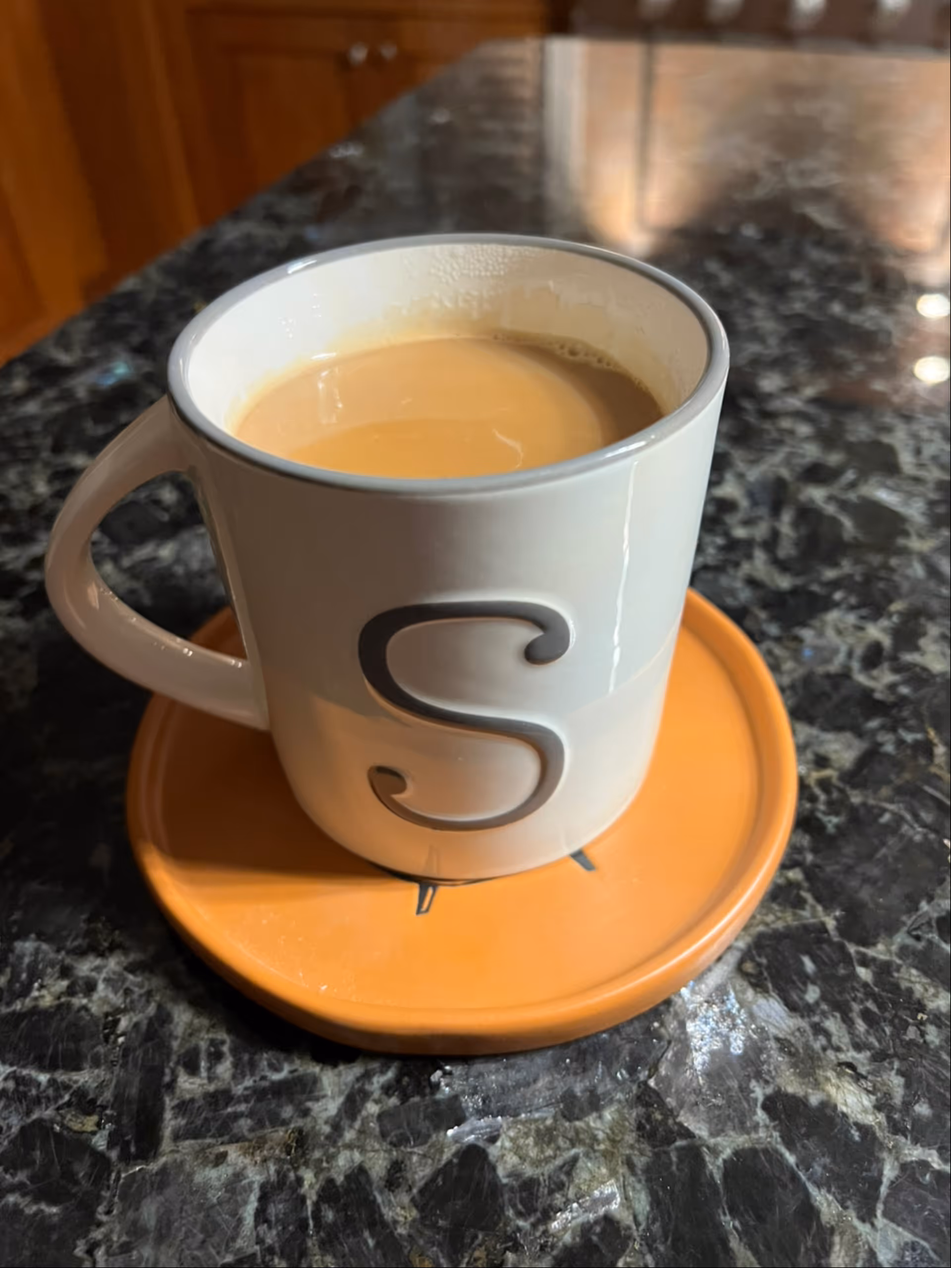 A white ceramic mug with the letter 'S' printed on it, filled with coffee or tea with cream, placed on an orange coaster on a dark granite countertop.