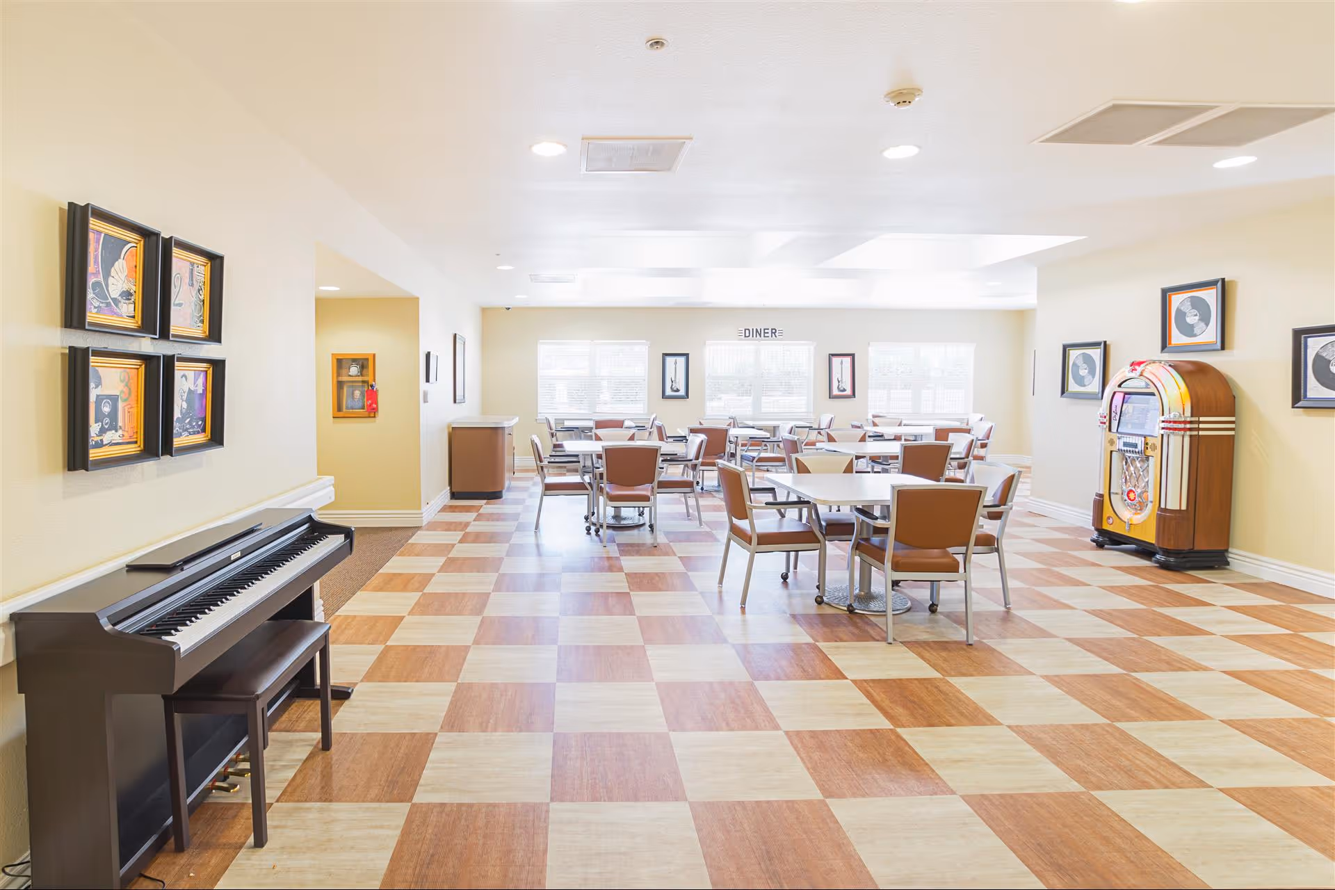 Bright dining area with checkered wood and beige flooring, several tables with chairs arranged for seating, a black piano with a bench on the left, framed artwork on the walls, and a vintage jukebox on the right side near the wall.