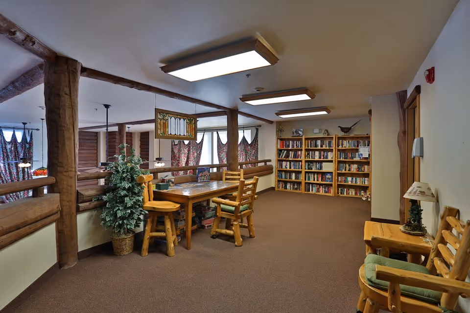 A cozy interior common area with wooden furniture including a table with chairs and a bookshelf filled with books. The space has rustic wooden beams, floral curtains on the windows, and warm lighting from ceiling fixtures. There is also a small lamp on a side table and a potted plant next to the table.