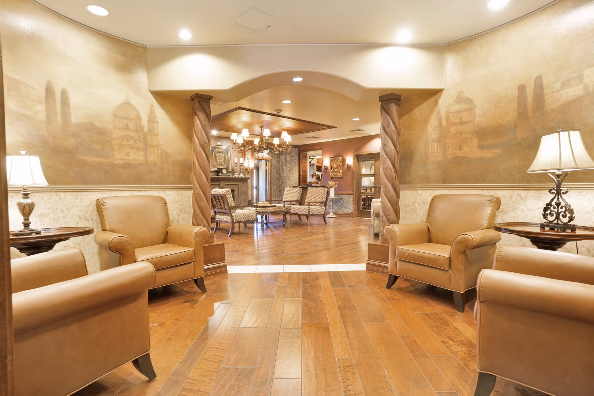 A warm and inviting living room area with tan leather armchairs arranged around wooden side tables with lamps. The room features wooden flooring, decorative columns, and a mural on the walls depicting architectural scenes. In the background, there is a cozy seating area with additional chairs and a chandelier hanging from the ceiling.