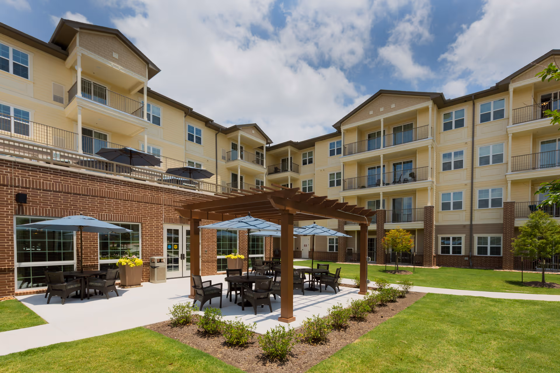 Outdoor courtyard area of a senior living facility with a pergola, tables, chairs, and umbrellas on a concrete patio. The building is a multi-story structure with balconies and large windows. The area is surrounded by well-maintained grass and small shrubs under a partly cloudy sky.