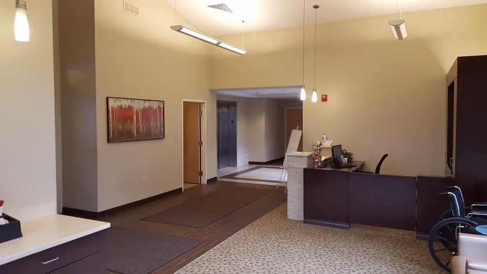 Interior view of a reception area in a skilled nursing facility. The image shows a reception desk with a computer and chair, a wheelchair nearby, pendant lights hanging from the ceiling, a hallway with elevators, and a piece of abstract artwork on the wall.