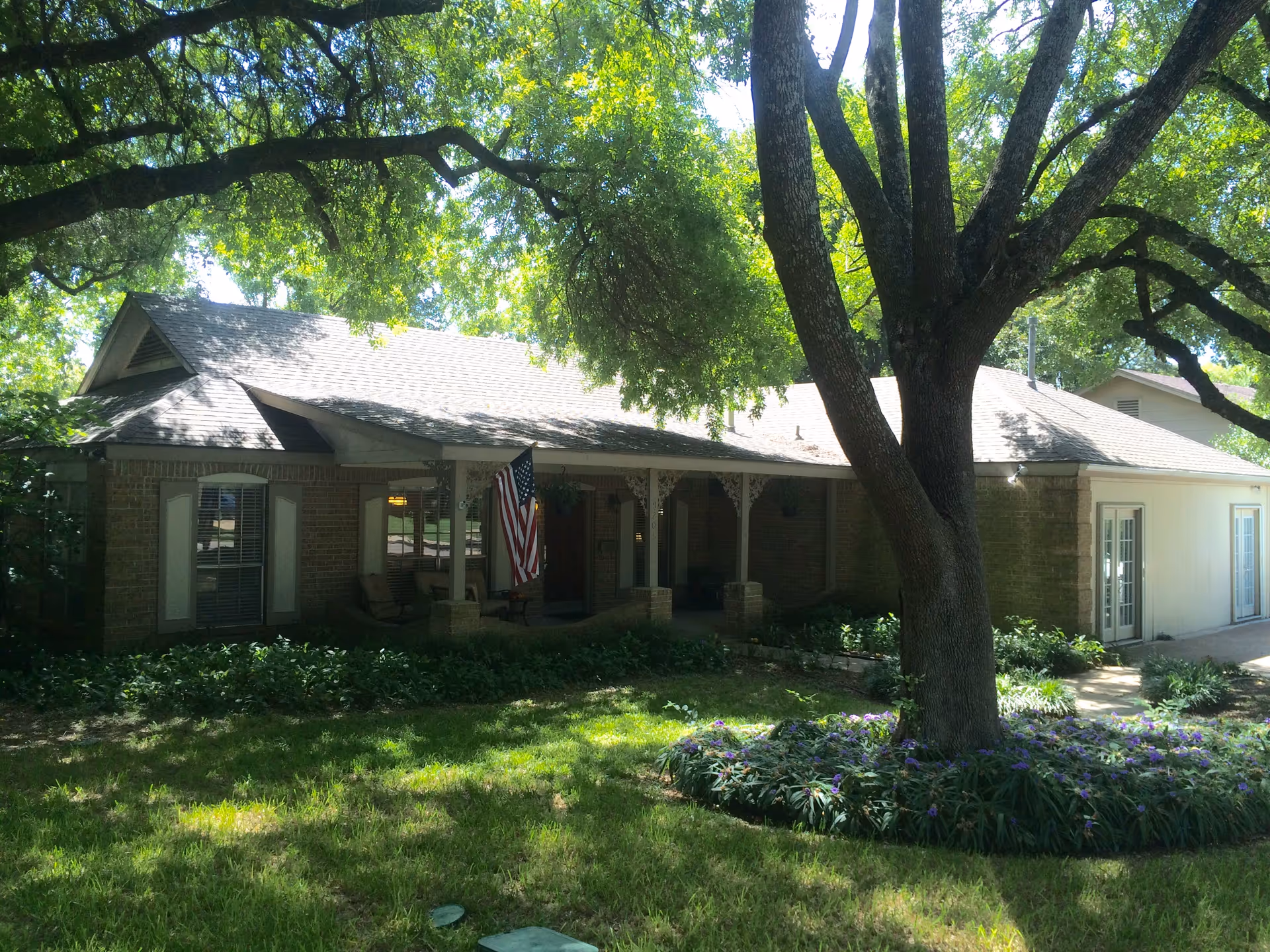 Single-story brick house with a covered front porch, an American flag, large shade trees and a grassy front yard.