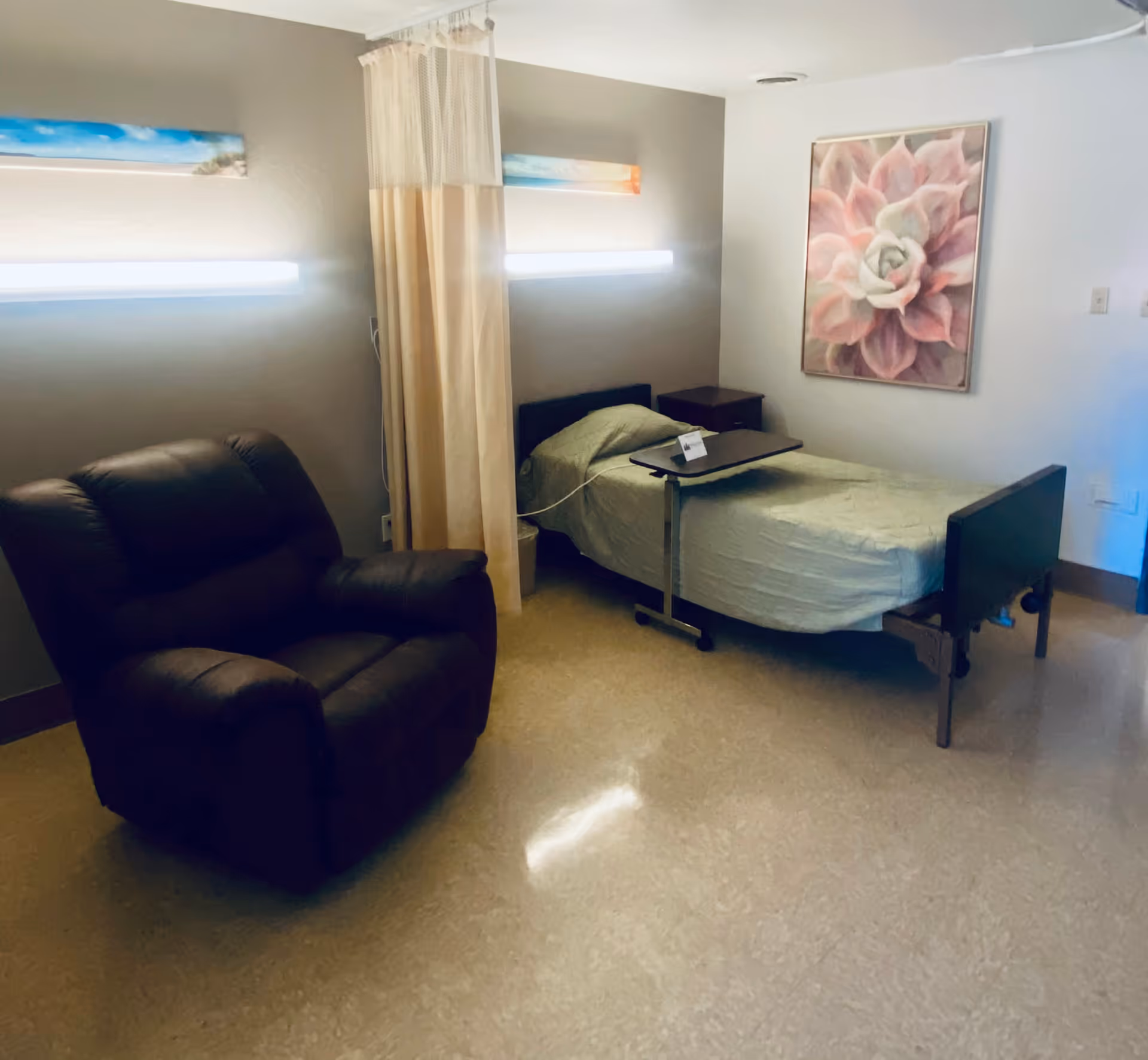 A senior living facility room with a single hospital-style bed covered in a light green blanket, a brown recliner chair, a small bedside table, and a floral painting on the wall. There is a privacy curtain partially drawn between the bed and the chair, and two horizontal wall lights above the bed.